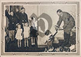 Recto carte postale ancienne avec Hitler fêtant Noel 1939 drapeau de la Hitlerjugend un enfant remets des fleurs à Hitler qui est surune marche avec bottes et brassard nazi et à gauche 5 enfants faisant le salut nazi 1939